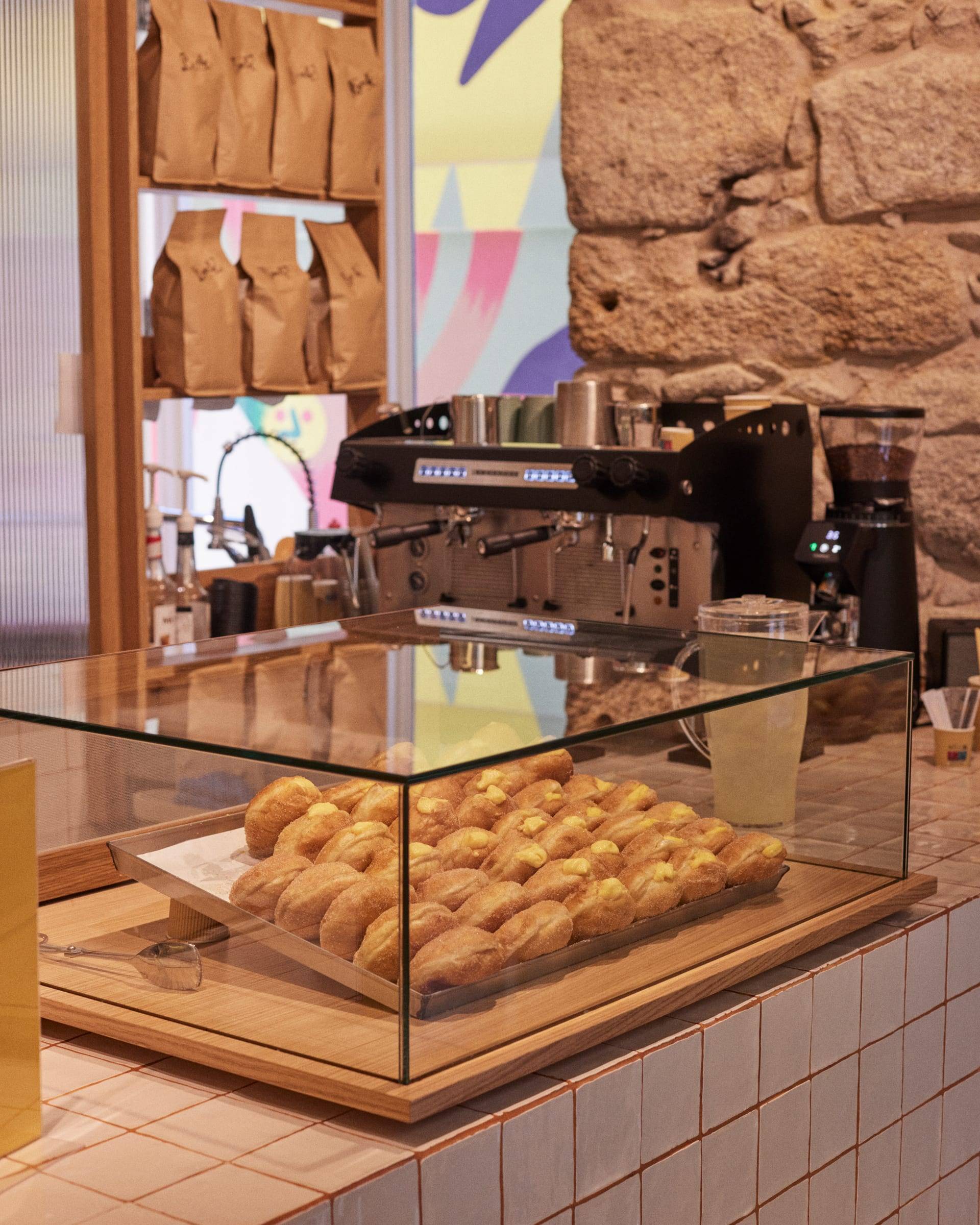 The shop counter: glass case of bolas, black espresso machine, exposed granite wall and kraft coffee bags