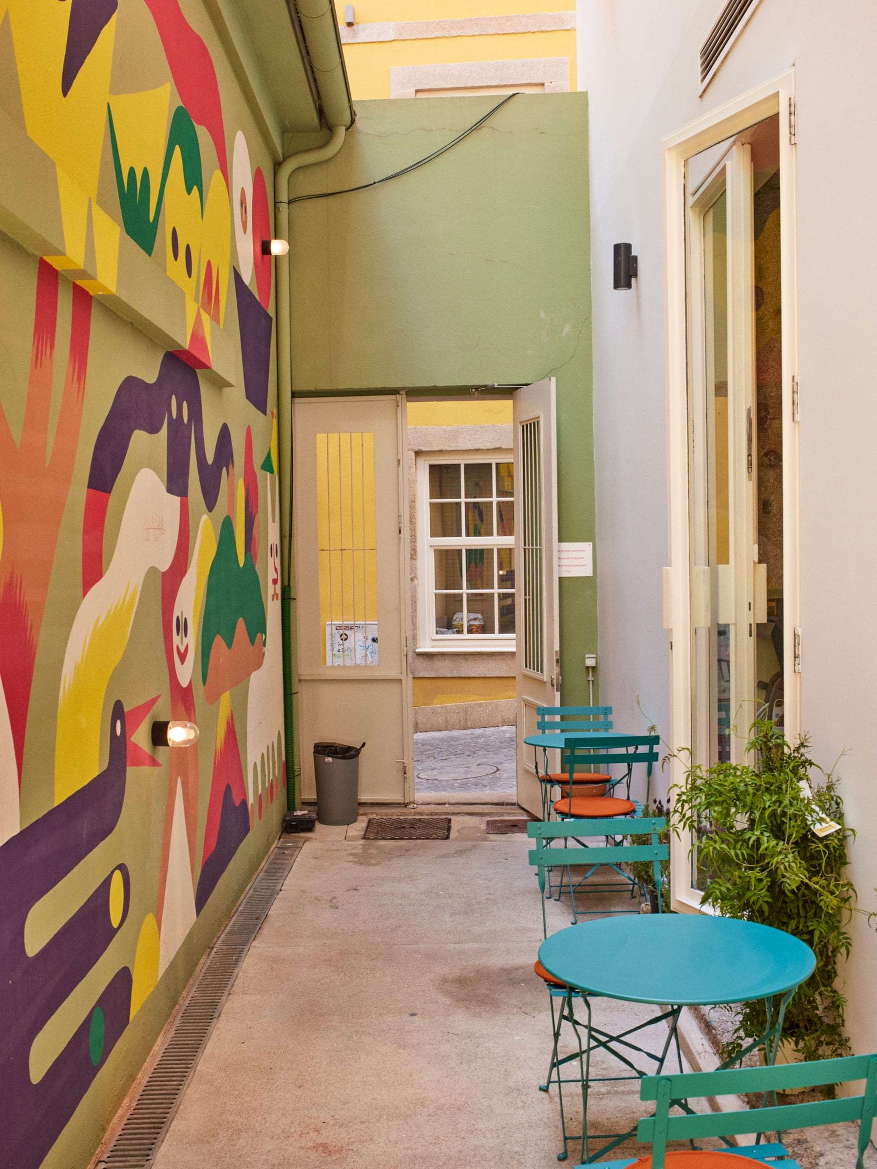 Side courtyard with a large mural of bold colourful shapes, turquoise bistro tables with orange seats and plants