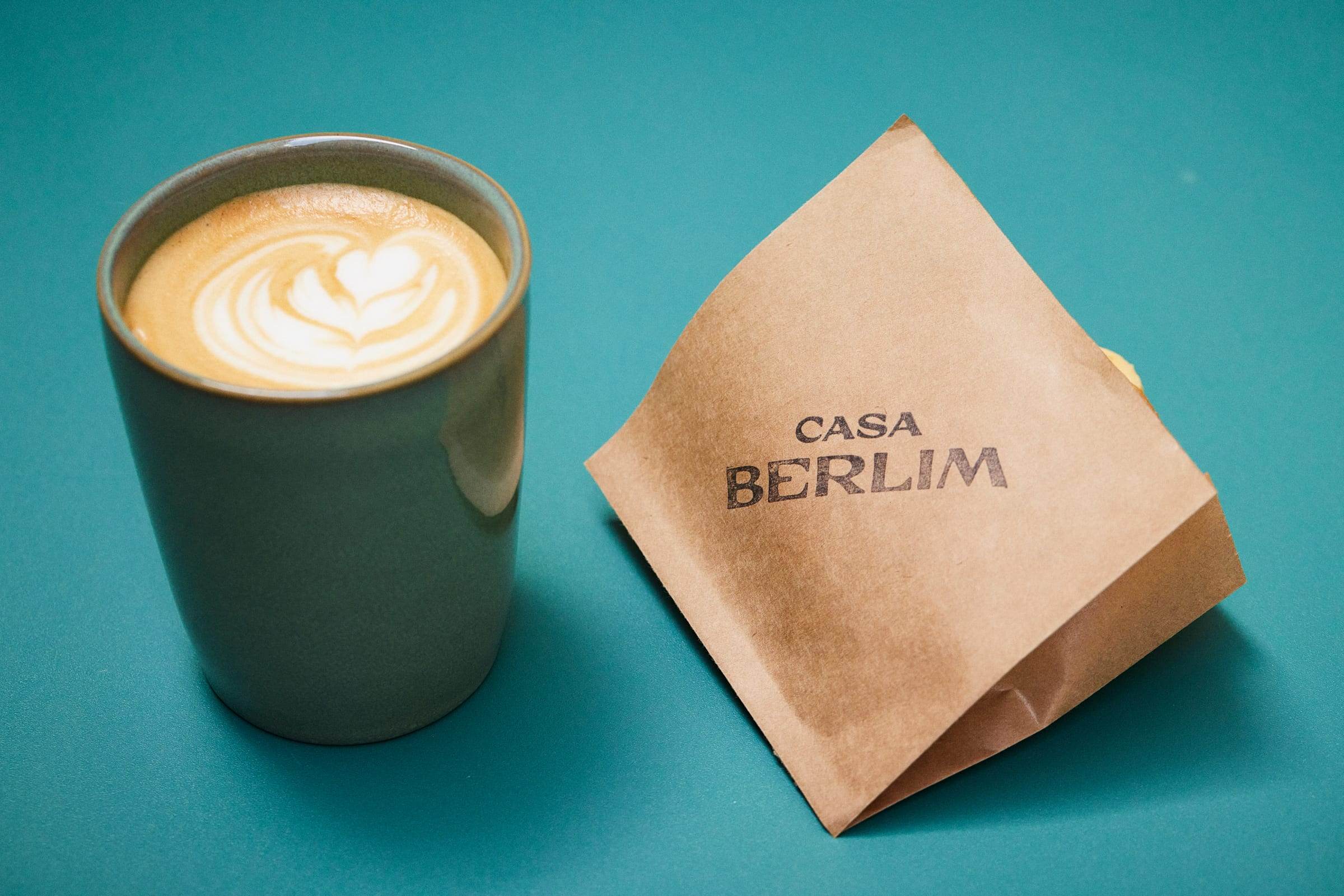 Green cup with latte art beside a kraft bag stamped CASA BERLIM, against a petrol-teal backdrop